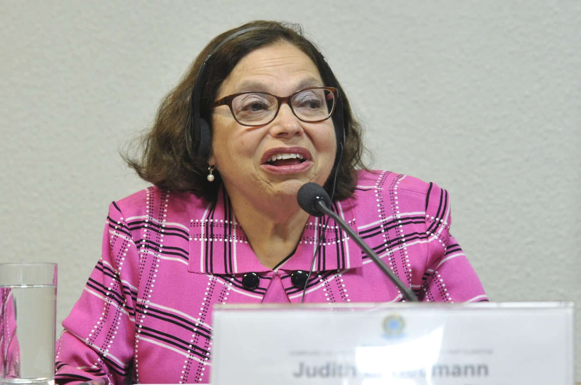 Judy Heummann, disability activist, dressed in a bright pink jacket, speaks into a microphone.