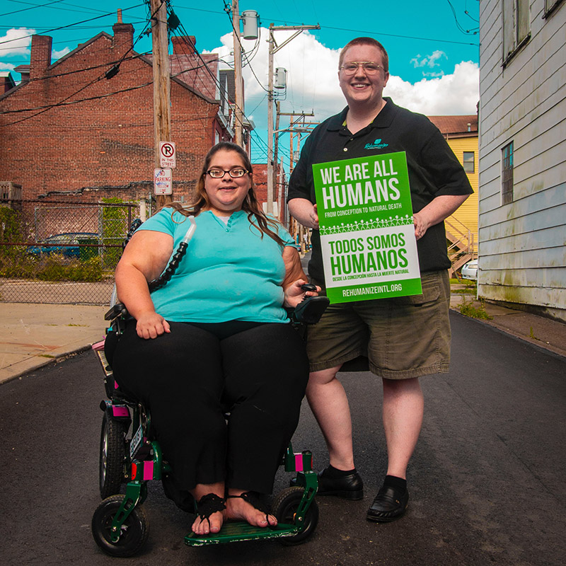 Woman using wheelchair and man standing holding sign that says we are all humans. 