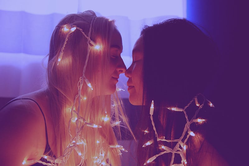 A blonde and brunette woman snuggle together under white lights. Be a brick in the wall of social movements for worldly problems.