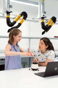 Two young women work in an engineering facility surrounded by robotic equipment. International Women's Day