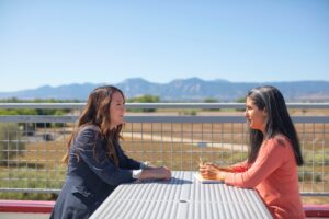 Two women seated at an outdoor table across from each other in front of a scenic mountain view. Greater good post.