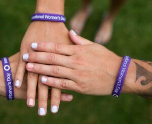 A stack of hands with purple International Women's Day bracelets on the wrists. How to be a feminist activist. Breaking down barriers for women and girls.