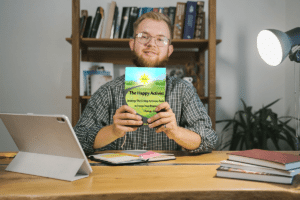 White man with glasses sits at a desk Holding The Happy Activist